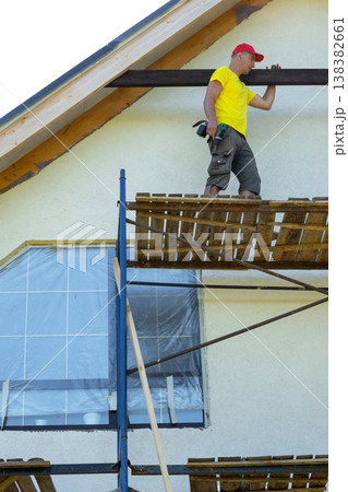 Worker on exterior scaffold fixes vertical plank to facade, yellow and white house, visible tools and handmade detail, renovation effort. Worker on exterior scaffold fixes vertical plank to facade, yellow and white house, visible tools and handmade detail, renovation effort. 138382661