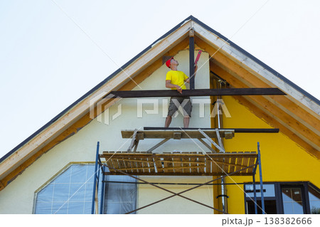 Yellow house with multiple scaffolds, craftsman places timber between beams, urban renovation, clear focus on manual labor, exterior upgrade ongoing. Yellow house with multiple scaffolds, craftsman places timber between beams, urban renovation, clear focus on manual labor, exterior upgrade ongoing. 138382666