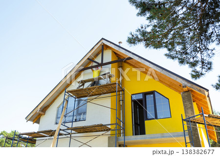 Man works on side of house installing decorative wooden design under roof slope, yellow gable and large window, DIY renovation on height. 138382677