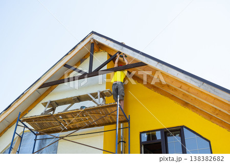 Craftsman in yellow shirt fits custom woodwork to gable peak, minimalist facade, working with measuring tape and toolbelt, DIY architecture focus. Craftsman in yellow shirt fits custom woodwork to gable peak, minimalist facade, working with measuring tape and toolbelt, DIY architecture focus. 138382682