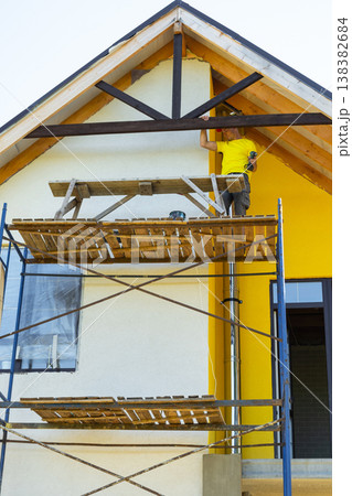 Man in helmet finalizes exterior painting of gable trim, high scaffold, modern yellow house, safety awareness, detail-oriented construction. 138382684