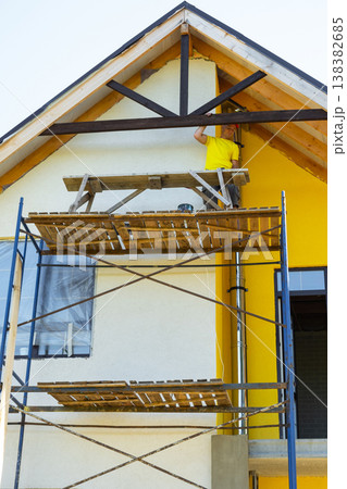 Builder inspects completed wooden gable design, stands on external scaffold, modern yellow facade, craftsmanship achievement, risk on height. Builder inspects completed wooden gable design, stands on external scaffold, modern yellow facade, craftsmanship achievement, risk on height. 138382685
