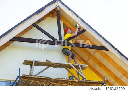 Male builder on scaffold reaches up to install vertical beam under the gable, minimalist yellow facade, DIY woodwork, expertise at height. 138382690