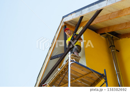 Worker sits on scaffold inspecting finished wood gable decor, modern yellow home, exterior architecture, emphasis on achievement and safety. Worker sits on scaffold inspecting finished wood gable decor, modern yellow home, exterior architecture, emphasis on achievement and safety. 138382692