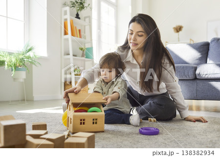 Mother and son playing wooden dollhouse game together at home, sitting on carpet in living room Mother and son playing wooden dollhouse game together at home, sitting on carpet in living room 138382934