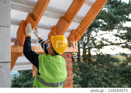 Builder in helmet attaches roof rafter to bearing wall during house construction, timber roof frame assembly, active construction phase. Builder in helmet attaches roof rafter to bearing wall during house construction, timber roof frame assembly, active construction phase. 138383075