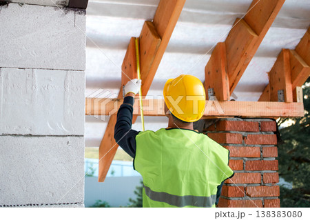 Male builder in vest checks alignment of rafters, fitting lag bolts for roof frame, visible brick support and insulation, house building. 138383080