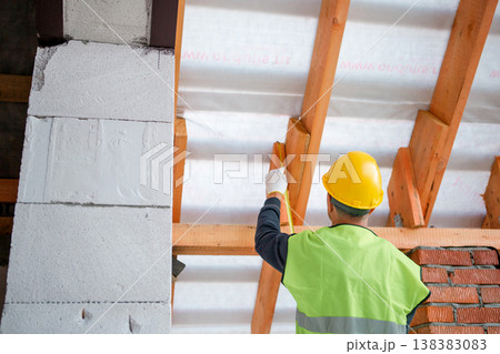 Worker using level tool adjusts wooden rafter during roofing, brick wall backdrop, house structure at roof installation stage. Worker using level tool adjusts wooden rafter during roofing, brick wall backdrop, house structure at roof installation stage. 138383083