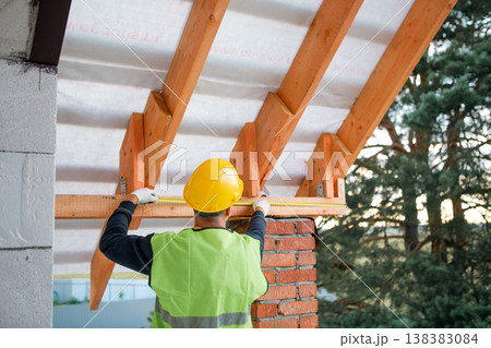 Male constructor secures ridge beam at roof peak, timber lags visible, advanced roofing in progress, house build frame close-up. 138383084
