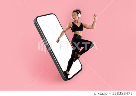 Sporty black woman with headphones jumping out of tilted large smartphone mockup Sporty black woman with headphones jumping out of tilted large smartphone mockup 138388475