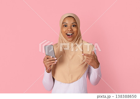 Surprised black woman in hijab reacting with excitement while pointing at smartphone Surprised black woman in hijab reacting with excitement while pointing at smartphone 138388650