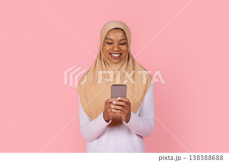 Young black Muslim woman in beige hijab smiling while looking at smartphone Young black Muslim woman in beige hijab smiling while looking at smartphone 138388688
