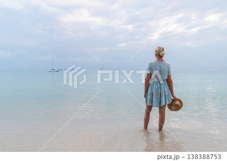 Young woman standing in shallow sea water holding straw hat and looking at calm ocean with boats on horizon Young woman standing in shallow sea water holding straw hat and looking at calm ocean with boats on horizon 138388753