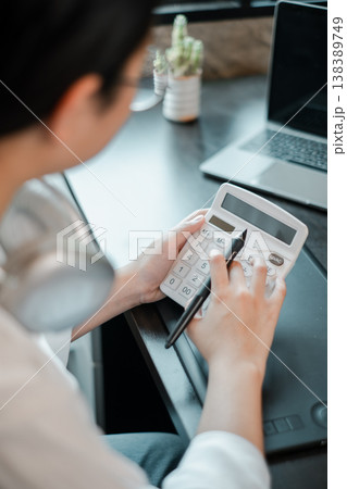 A person using a calculator at a desk with a laptop and plant, focusing on financial tasks in a modern office setting. A person using a calculator at a desk with a laptop and plant, focusing on financial tasks in a modern office setting. 138389749