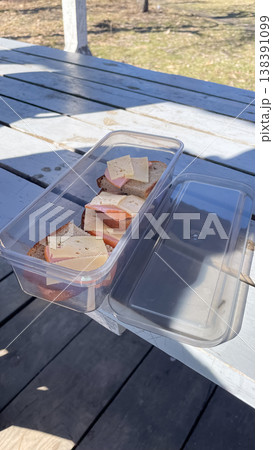 Plastic lunch box with fresh ham and cheese sandwiches on a rustic wooden table during a spring picnic 138391099