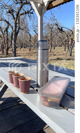Stainless steel thermos with paper cups and a lunch box on a wooden table in a sunny spring orchard 138391106