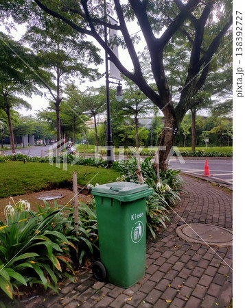 A green trash bin stands neatly on a clean sidewalk, surrounded by lush trees and urban calm. A green trash bin stands neatly on a clean sidewalk, surrounded by lush trees and urban calm. 138392727