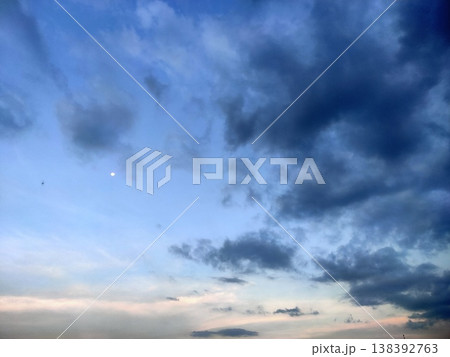 A calm sky at dusk with scattered clouds and a soft moon glowing in the serene blue horizon. 138392763