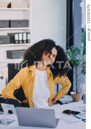 Businesswoman experiencing discomfort and pain in her lower back while sitting at her desk, working on a laptop in a modern office environment, highlighting work-related health issues and stress 138392976