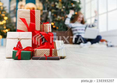 Winter holidays background. Christmas gift boxes band with red ribbons closeup at decorated xmas tree and happy young girl backdrop. Selective focus, copy space Winter holidays background. Christmas gift boxes band with red ribbons closeup at decorated xmas tree and happy young girl backdrop. Selective focus, copy space 138393207