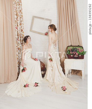 Wedding dress fashion. Two beautiful young brides in vintage wedding dresses indoors. Rustic wedding dresses at models. Girls in champaigne color dresses in decorated interior with flowers, high key. 138393282