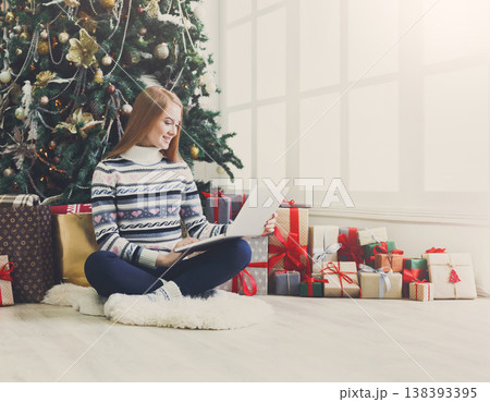 Young woman shopping online on laptop in cozy christmas interior. Girl in casual sitting under decorated pine tree among lots of wrapped presents. Preparing to xmas, bying on winter sales. Copy space 138393395