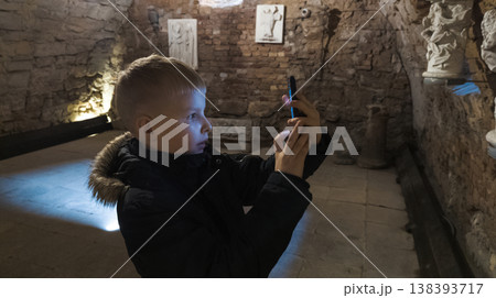 Boy photographing museum exhibits in an underground vault, exploring history with a smartphone. Low light travel scene, child curiosity, education and digital exploration. Boy photographing museum exhibits in an underground vault, exploring history with a smartphone. Low light travel scene, child curiosity, education and digital exploration. 138393717