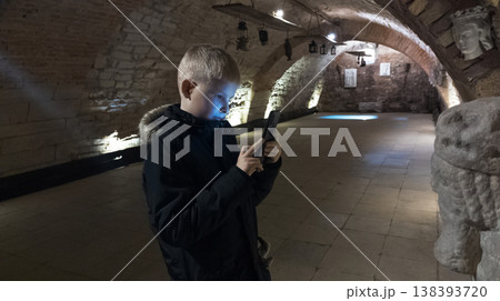 Boy photographing museum exhibits in an underground vault, exploring history with a smartphone. Low light travel scene, child curiosity, education and digital exploration. Boy photographing museum exhibits in an underground vault, exploring history with a smartphone. Low light travel scene, child curiosity, education and digital exploration. 138393720