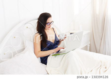 Middle aged woman with laptop in white bed. Woman in eyeglasses looks into gadget pc. Morning coffee. Beautiful woman in blue nightwear at bed surfing internet at her computer. High key. 138394126