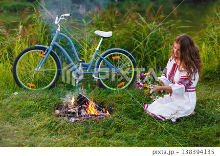 Woman in Folk costume with bicycle. Young attractive woman at riverside having rest with bicycle in national ethnic ukrainian folk traditional dress. Girl looking at circlet of flowers near bonfire. 138394135