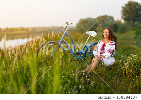 Woman in Folk costume with bicycle. Young attractive woman at the riverside having rest with bicycle in national ethnic ukrainian folk traditional costume or dress. 138394137