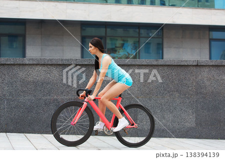 Sexy girl with bicycle. Young slim sexy sporty woman in blue shorts and white snickers long-haired, sensual riding at pink fix bicycle in urban city environment. Soft toned. 138394139