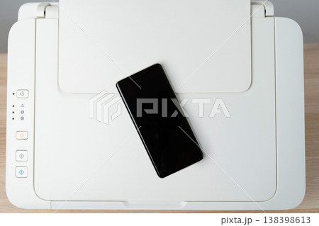 Overhead view of a blank smartphone resting on a white multifunction printer on a desk, illustrating office technology, mobile connectivity, printing, scanning and minimalist workspace productivity. Overhead view of a blank smartphone resting on a white multifunction printer on a desk, illustrating office technology, mobile connectivity, printing, scanning and minimalist workspace productivity. 138398613