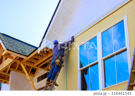 Worker is using ladder to place siding on new building at construction site. Worker is using ladder to place siding on new building at construction site. 138401545