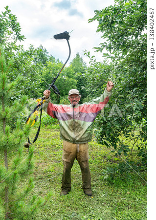Elderly man holds electric string trimmer proudly in green orchard Elderly man holds electric string trimmer proudly in green orchard 138402487
