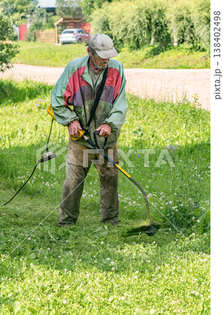 Elderly man trims grass with electric trimmer in his yard on a sunny day Elderly man trims grass with electric trimmer in his yard on a sunny day 138402498