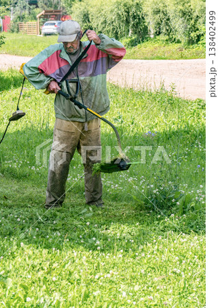 Elderly man mows grass with an electric trimmer in front of his house in residential area Elderly man mows grass with an electric trimmer in front of his house in residential area 138402499
