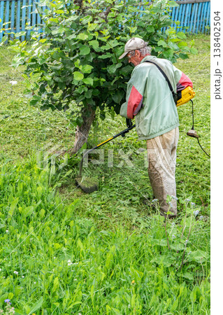 Elderly man mows grass with electric trimmer in front of his home 138402504