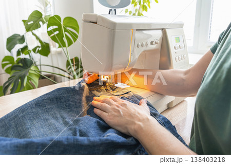 Hands operating a sewing machine, stitching blue denim fabric of jeans, repairing clothing. Person sewing denim jeans on a sewing machine 138403218