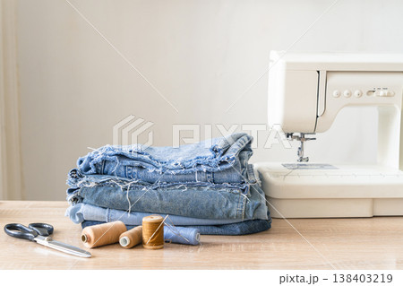 Stacked denim fabric, scissors, and thread next to a sewing machine, preparing for clothing repair and upcycling. Reusing things. Environmental awareness. Eco-friendly lifestyle Stacked denim fabric, scissors, and thread next to a sewing machine, preparing for clothing repair and upcycling. Reusing things. Environmental awareness. Eco-friendly lifestyle 138403219