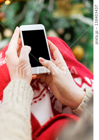 Christmas online shopping. Female buyer makes order at screen of smartphone with copy space. Woman buys presents for xmas eve, sits at home near pine tree with shining lights. Winter holidays sales 138403437