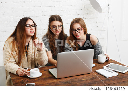 Three young women sit at a wooden table, focused on a laptop. They are discussing something important while sipping coffee. Notebooks and a tablet are on the table as they collaborate. 138403439