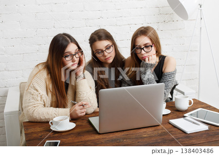 Three young women are sitting around a table. They are focused on the laptop screen while drinking coffee. The room has a bright atmosphere with white walls and simple decor. 138403486