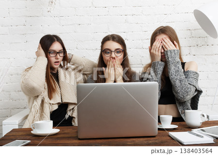 Three friends gather around a laptop in a cafe. They show expressions of surprise and disbelief while drinking coffee. The moment captures their shared experience and reactions. 138403564