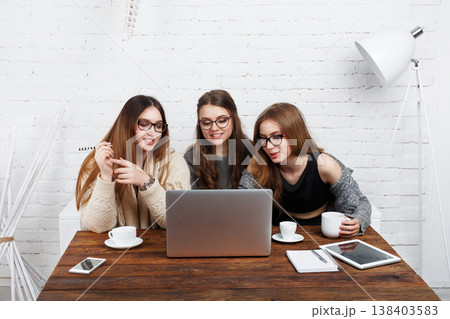 Three friends gather around a laptop on a wooden table. They share ideas while drinking coffee. The setting has bright walls and modern decor. Notes are scattered on the table. 138403583