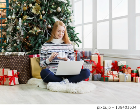 Young woman with coffee shopping online on laptop in christmas interior. Girl sitting under decorated fir tree among lots of wrapped presents. Preparing to xmas, bying on winter sales, copy space Young woman with coffee shopping online on laptop in christmas interior. Girl sitting under decorated fir tree among lots of wrapped presents. Preparing to xmas, bying on winter sales, copy space 138403634