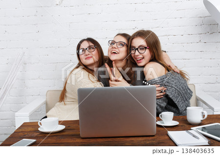 Three friends gather around a laptop, smiling and embracing each other. They enjoy their time together while working in a bright room with coffee cups on the table. Three friends gather around a laptop, smiling and embracing each other. They enjoy their time together while working in a bright room with coffee cups on the table. 138403635
