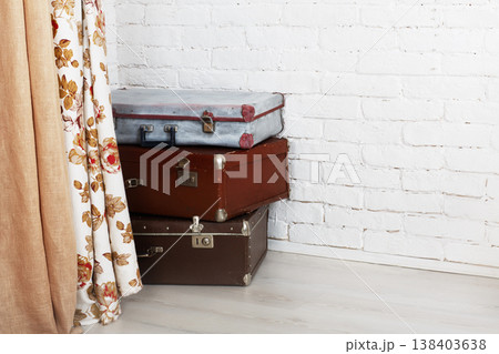 Three vintage suitcases are stacked in a corner against a white brick wall. A floral curtain hangs beside the suitcases, adding color to the bright space. 138403638