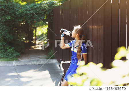 Female sportsman cyclist drinks water from the bottle at the race. Riding racing bicycle. Break, stop at road. Woman cycling on countryside summer road or highway. Triathlon or cycling competition. Female sportsman cyclist drinks water from the bottle at the race. Riding racing bicycle. Break, stop at road. Woman cycling on countryside summer road or highway. Triathlon or cycling competition. 138403670