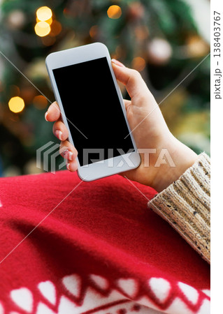 Christmas online shopping. Female buyer makes order at screen of smartphone with copy space. Woman buys presents for xmas eve, sits at home near pine tree with shining lights. Winter holidays sales 138403767